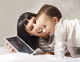 A mom in a striped shirt and a toddler in polka dots smile at a tablet screen as they video chat with grandma and grandpa.