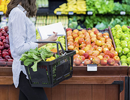 Woman in the produce department of a grocery store with a shopping basket and a grocery list.